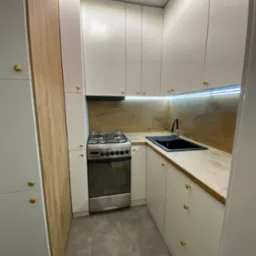 Niewielka kuchnia z białymi szafkami, złotymi uchwytami, drewnianym blatem i panelem ściennym, oświetleniem LED pod szafkami wiszącymi oraz czarnym zlewozmywakiem i srebrną kuchenką.