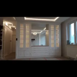 Biała zabudowa meblowa z przeszklonymi szafkami i lustrem w centralnej części, oświetlenie LED podkreśla detale, widoczne elementy wykończenia wnętrza.