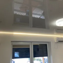 Biały, błyszczący sufit napinany z wbudowanym oświetleniem LED i odbiciem okna, widoczne klimatyzatory na ścianie.