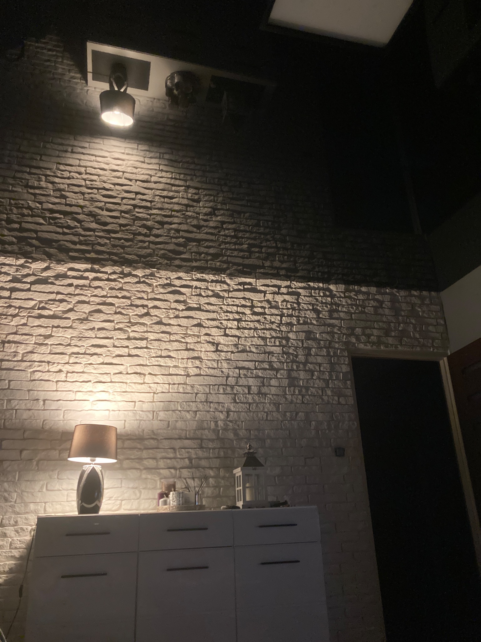 Ściana z cegły dekoracyjnej oświetlona lampą sufitową i lampką nocną na komodzie w ciemnym wnętrzu.