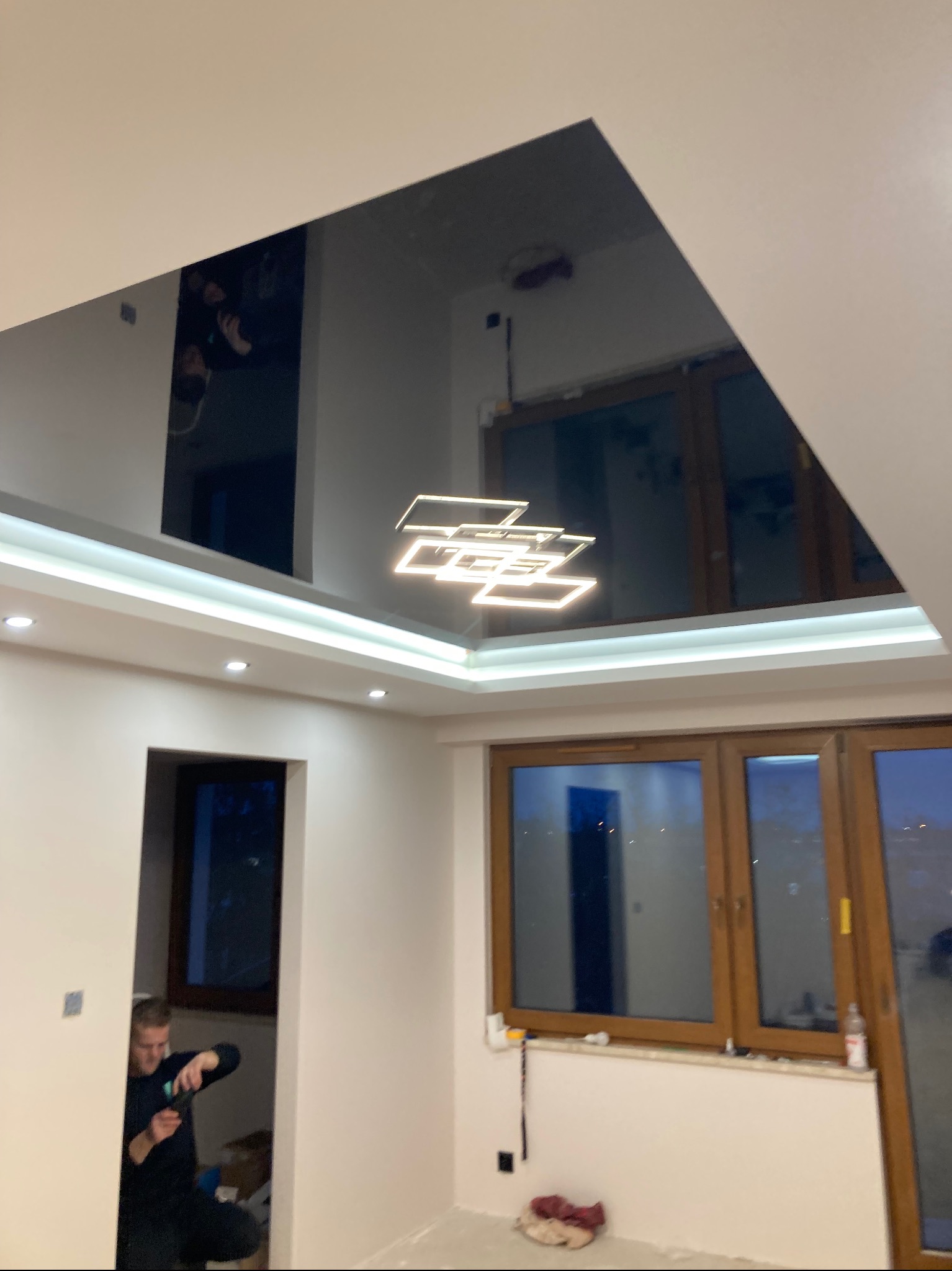 Błyszczący, czarny sufit napinany z nowoczesnym oświetleniem LED w kształcie kwadratów, odbijający fragment pokoju i sylwetkę osoby; widoczny pracownik w trakcie prac wykończeniowych.