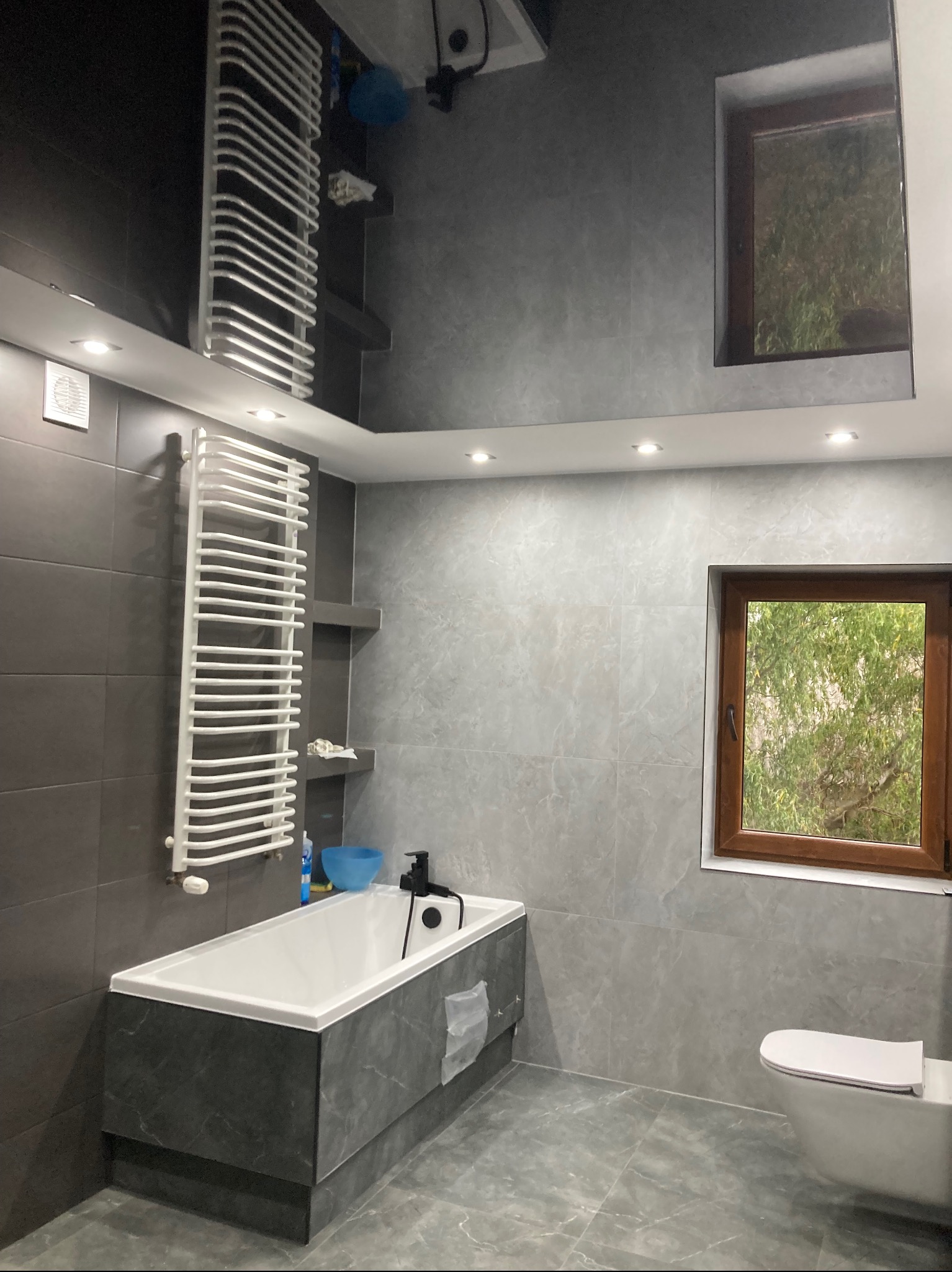Nowoczesna łazienka w szarościach z białym grzejnikiem drabinkowym, wanną i toaletą, okno z widokiem na zieleń, błyszczący sufit z oświetleniem punktowym.