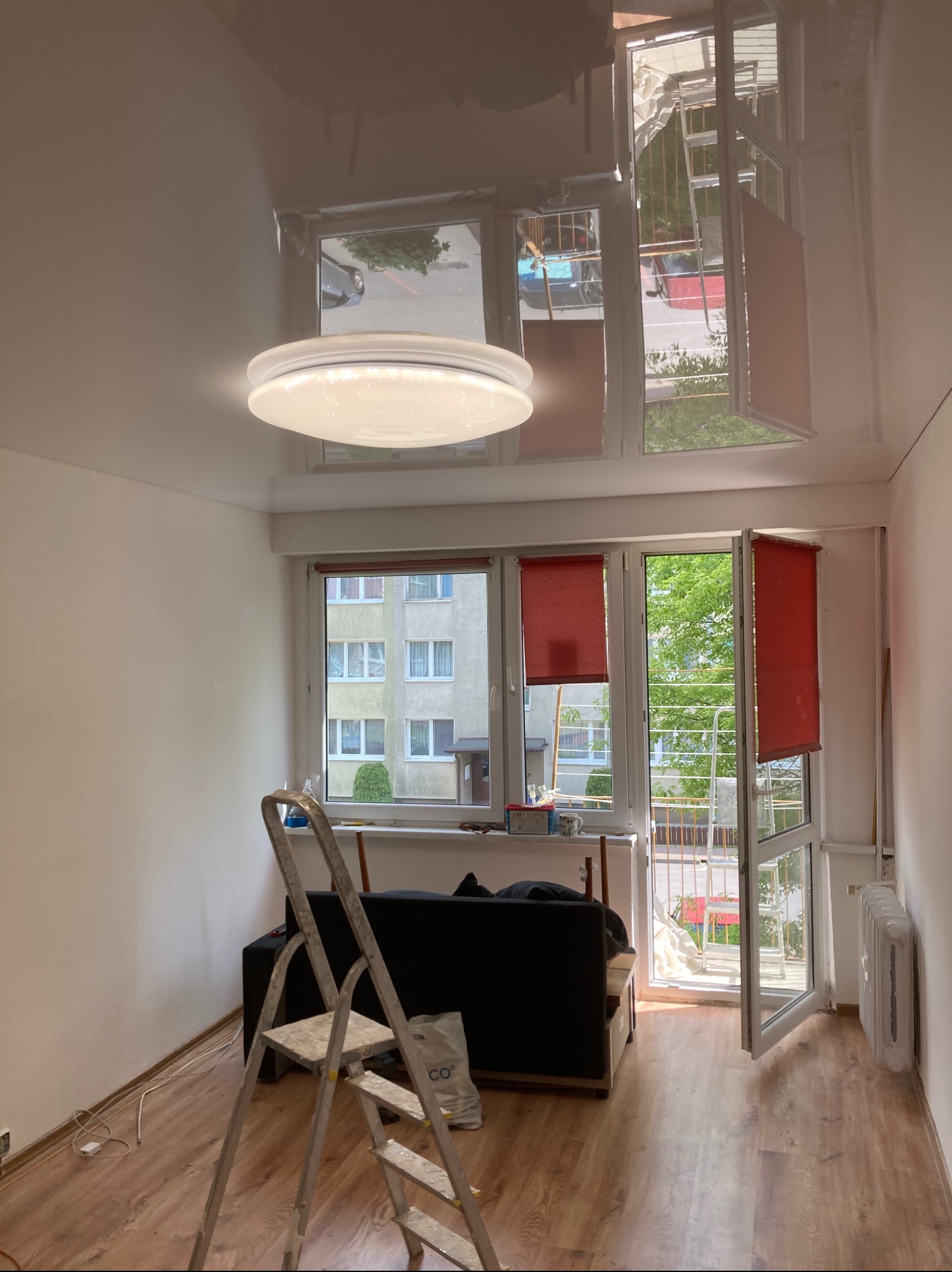Błyszczący sufit napinany w pokoju z oknami balkonowymi, widoczna drabina i lampa sufitowa