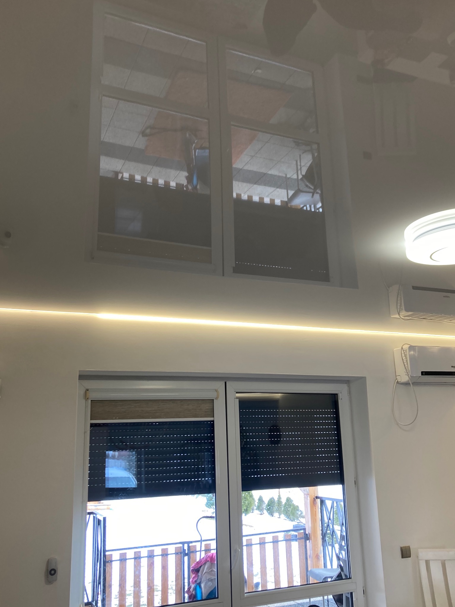 Biały, błyszczący sufit napinany z wbudowanym oświetleniem LED i odbiciem okna, widoczne klimatyzatory na ścianie.