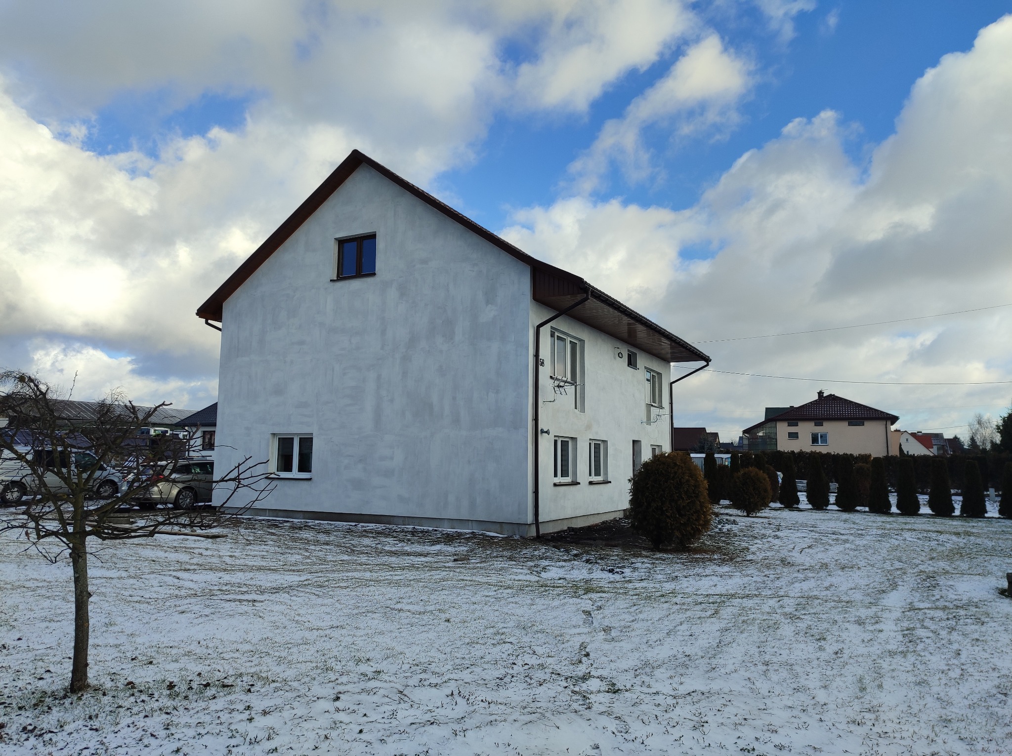 Dwukondygnacyjny dom z elewacją w odcieniach szarości, widoczny z boku w zimowej scenerii, z częściowo ośnieżonym trawnikiem i nagim drzewem na pierwszym planie.