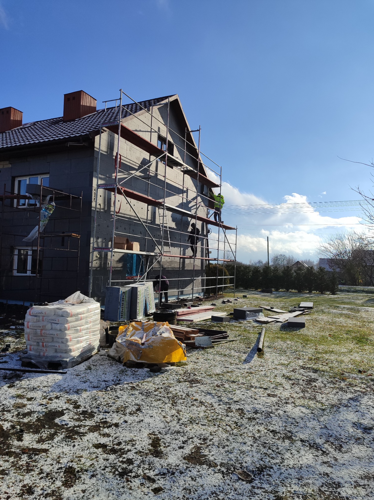 Dom jednorodzinny w Rejowcu w trakcie prac związanych z ociepleniem elewacji, widoczne rusztowania, materiały izolacyjne i pracownicy na tle błękitnego nieba z chmurami.