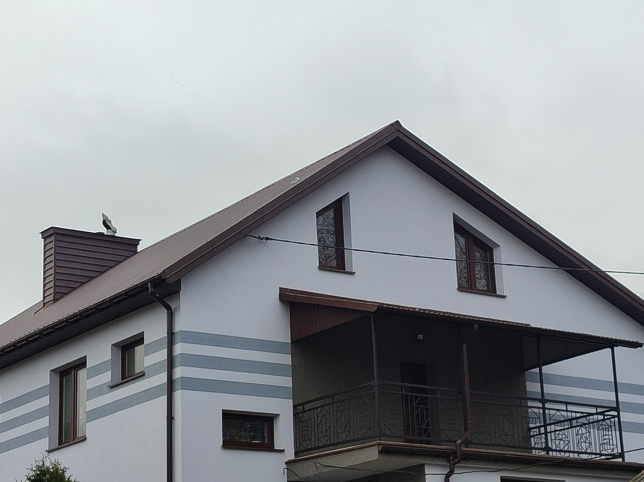 Dwupiętrowy dom z białymi ścianami i brązowym dachem, z widocznymi szarymi pasami dekoracyjnymi na elewacji i balkonem z metalową balustradą.