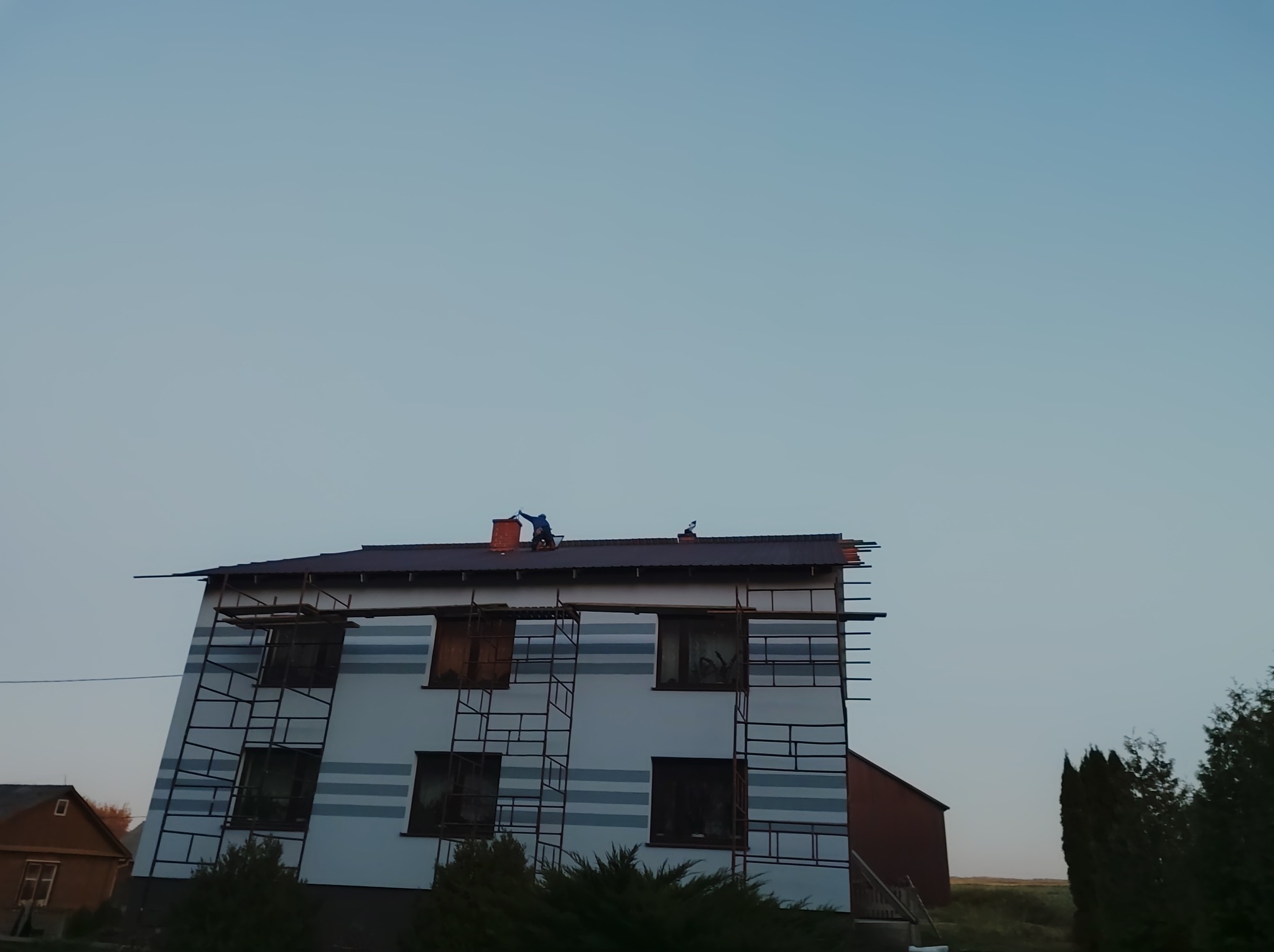 Dwupiętrowy dom w trakcie prac wykończeniowych elewacji, z widocznymi rusztowaniami, poziomymi pasami na ścianie i dwoma pracownikami na dachu w oddali.