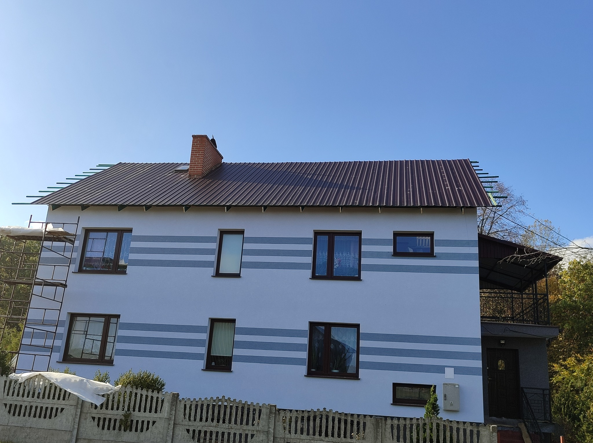 Dwupiętrowy dom z szarą elewacją w poziome pasy, brązowym dachem z metalu i ceglanym kominem, w trakcie montażu obróbek blacharskich dachu, widoczny fragment rusztowania.