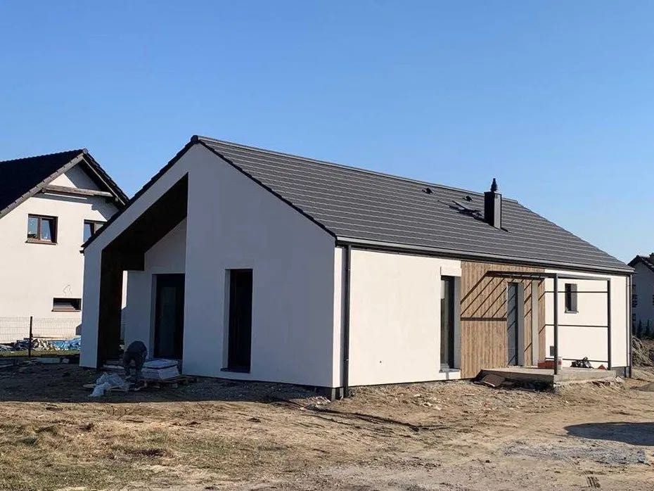 Nowoczesny dom jednorodzinny z białą elewacją i elementami drewnianymi, widoczne prace wykończeniowe wokół budynku, błękitne niebo.