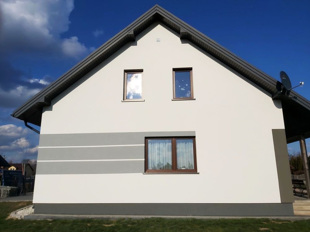 Wykończona elewacja domu jednorodzinnego z białym tynkiem, szarymi pasami i ciemną podbitką dachową, okna z brązowymi ramami, widoczne odbicie chmur.
