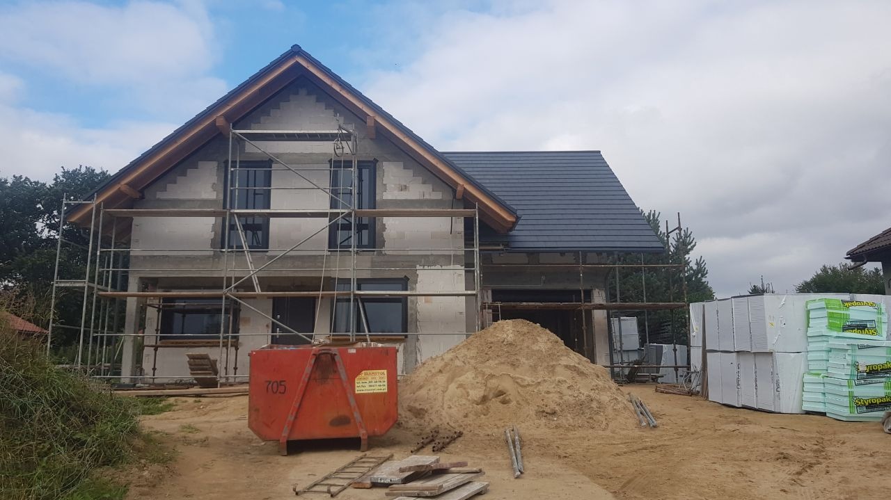 Dom w trakcie budowy z zamontowanymi oknami, częściowo otynkowany, z rusztowaniami, stertą piasku i kontenerem na gruz na pierwszym planie. Widoczne paczki styropianu. Dach pokryty ciemną dachówką.