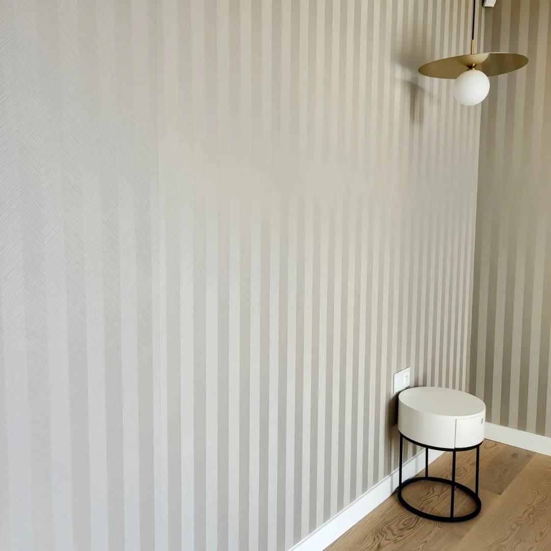 Wnętrze pokoju po remoncie: ściany w beżowo-białe pionowe pasy, okrągły stolik nocny z szufladą, lampa sufitowa z białym kloszem i złotym elementem.