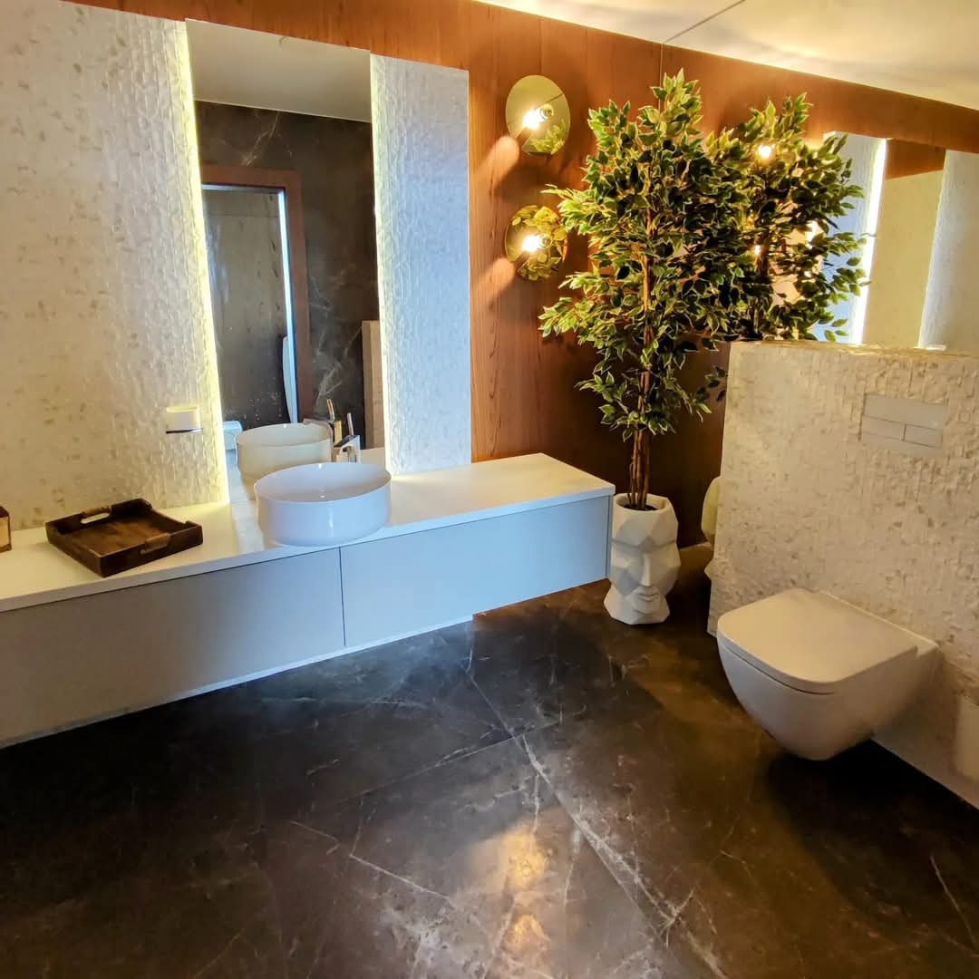 Nowoczesna łazienka z marmurową podłogą, białą umywalką nablatową i toaletą podwieszaną, oświetlona złotymi lampami ściennymi, z dekoracyjną rośliną w doniczce w kształcie twarzy.