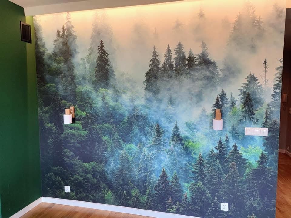Fototapeta z motywem mglistego lasu iglastego na ścianie w pokoju, drewniana podłoga, zielona ściana po lewej stronie, widoczne gniazdka elektryczne i włączniki światła.