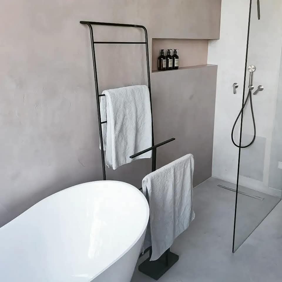 Minimalistyczna łazienka z wolnostojącą wanną, czarnym wieszakiem na ręczniki w formie drabiny, szklaną kabiną prysznicową i butelkami kosmetyków w niszach ściennych.