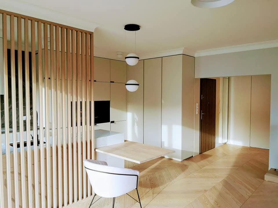 Jasne wnętrze z drewnianą podłogą w jodełkę, drewnianą ażurową ścianką działową, białym krzesłem i wiszącymi kulistymi lampami, minimalistyczna zabudowa kuchenna w tle.