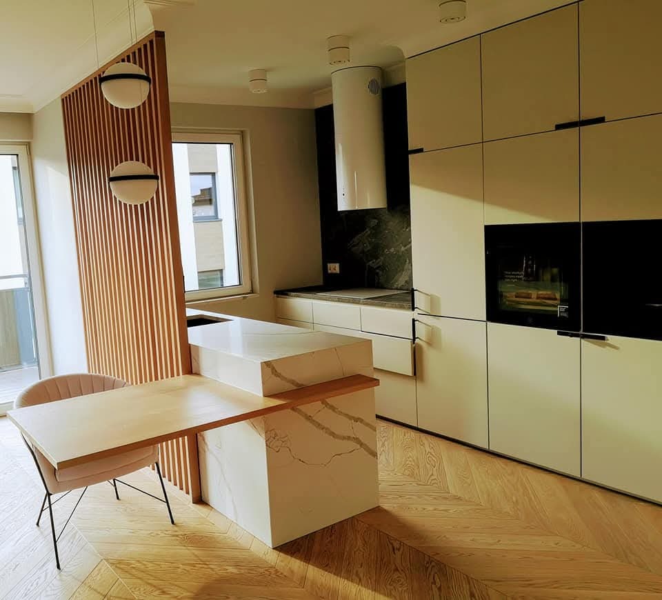 Jasna kuchnia z drewnianą podłogą w jodełkę, wyspą z blatem i marmurowym wykończeniem, oddzielona od salonu drewnianą ścianką lamelową z okrągłymi lampami wiszącymi.