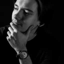 Czarno-biały portret młodego mężczyzny z zamyśloną miną, ręka podparta pod brodą, widoczny zegarek na nadgarstku.