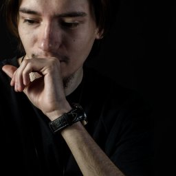 Kot Studio Fotograficzno-Graficzne Oliwia Kot - Portret zamyślonego mężczyzny z ręką przy ustach, w czarnej koszulce i z zegarkiem na skórzanym pasku na ciemnym tle.