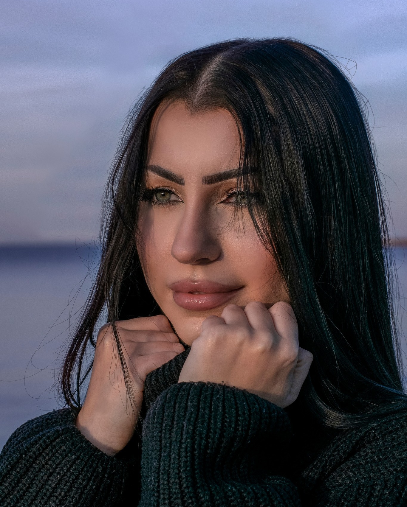 Portret kobiety z długimi, czarnymi włosami, opierającej brodę na dłoniach w ciemnym swetrze, z zamyślonym spojrzeniem na tle rozmytego krajobrazu wodnego o zachodzie słońca.