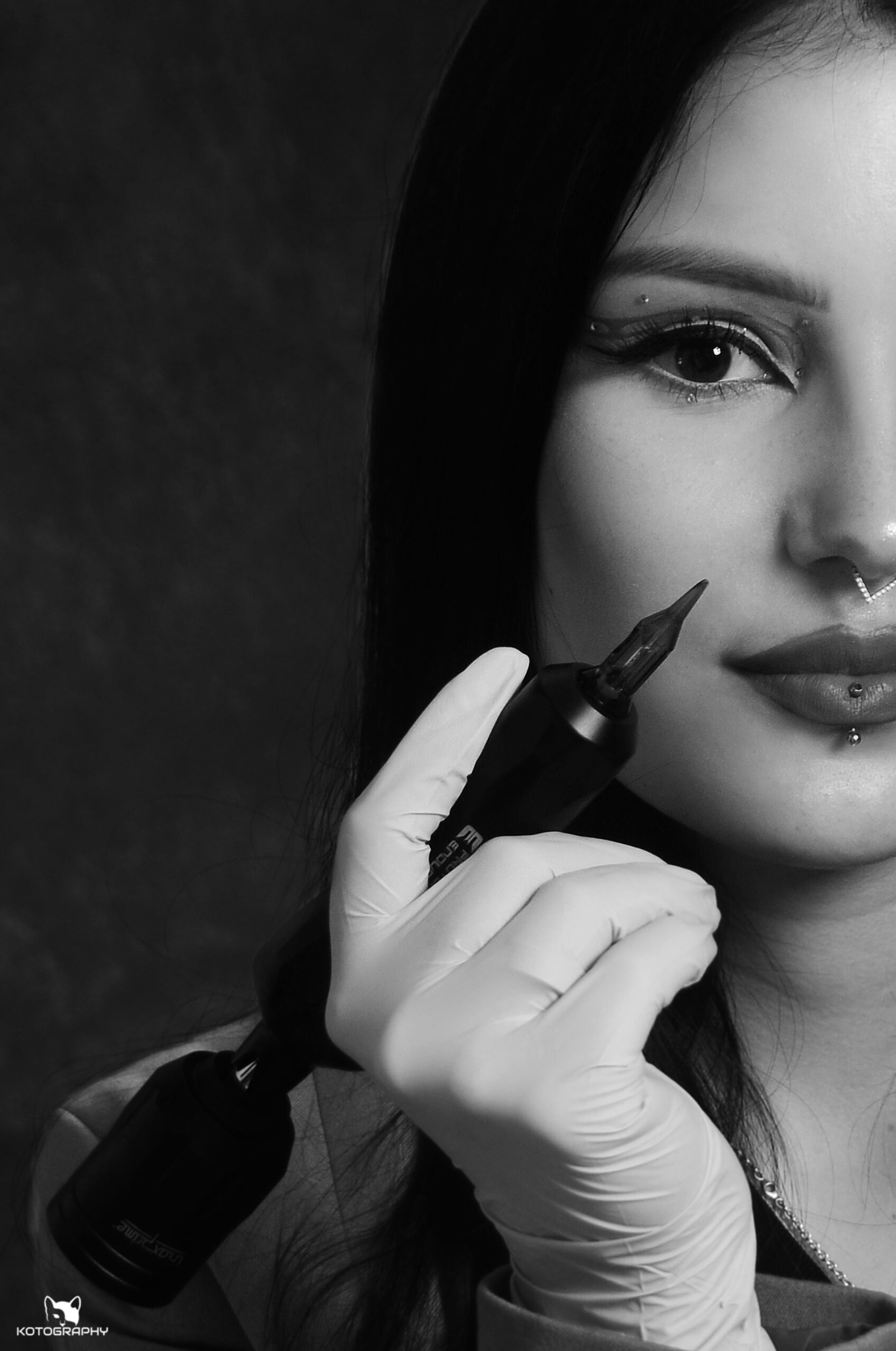 Czarno-białe zbliżenie na twarz kobiety z piercingiem i ozdobnym makijażem oczu, trzymającej w rękawicy jednorazowej maszynkę do tatuażu.