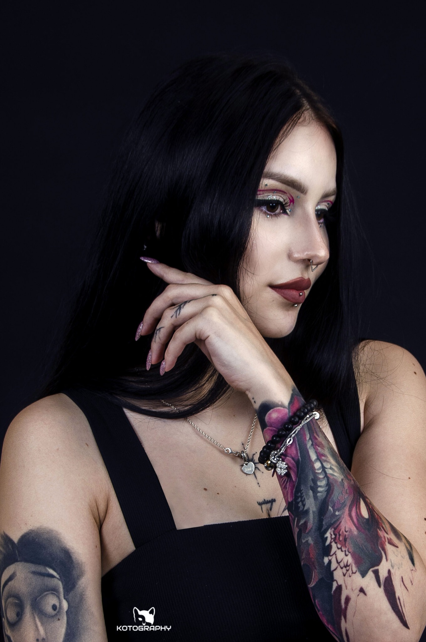 Portret kobiety z ciemnymi włosami, wyrazistym makijażem oczu z brokatem, piercingiem w nosie i ustach, tatuażami na rękach oraz manicure z brokatem, na czarnym tle.