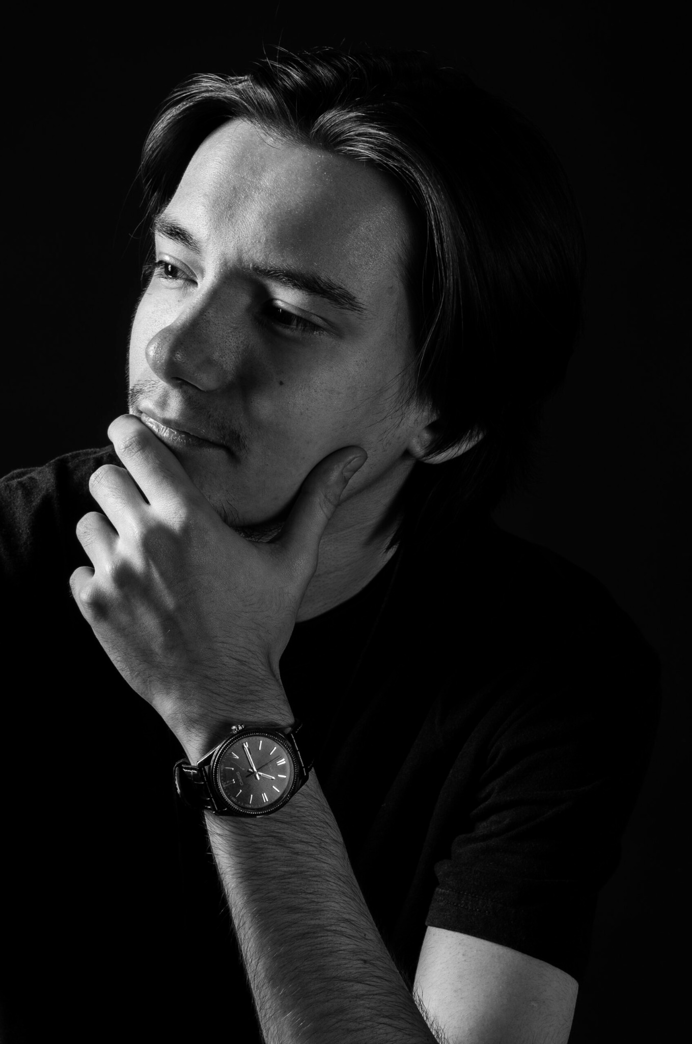 Czarno-biały portret młodego mężczyzny z zamyśloną miną, ręka podparta pod brodą, widoczny zegarek na nadgarstku.