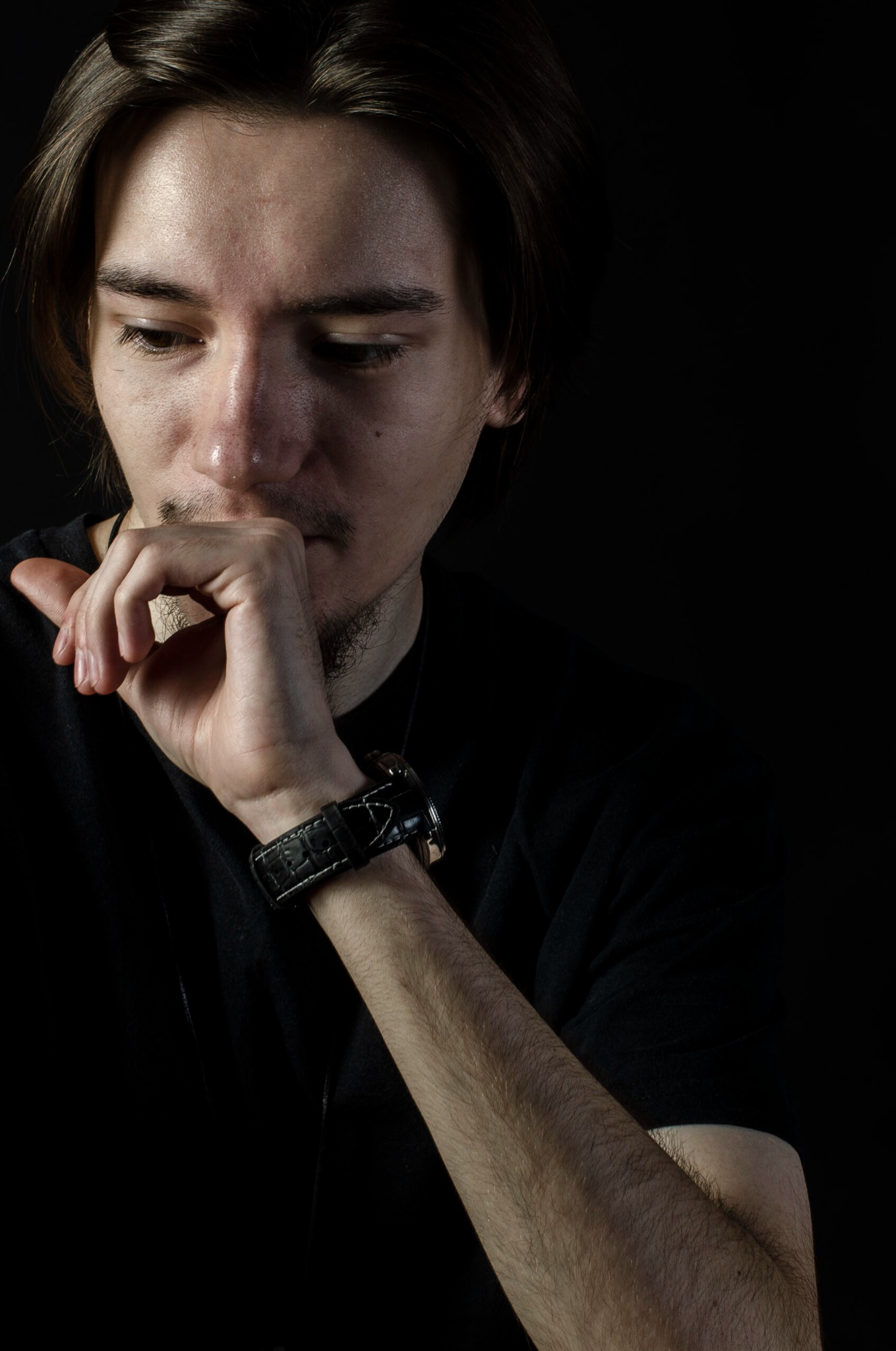 Portret zamyślonego mężczyzny z ręką przy ustach, w czarnej koszulce i z zegarkiem na skórzanym pasku na ciemnym tle.