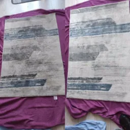 Dwa identyczne dywany z abstrakcyjnym wzorem w odcieniach szarości i błękitu, leżące na fioletowym kocu. Jeden dywan ma widoczną małą białą metkę.