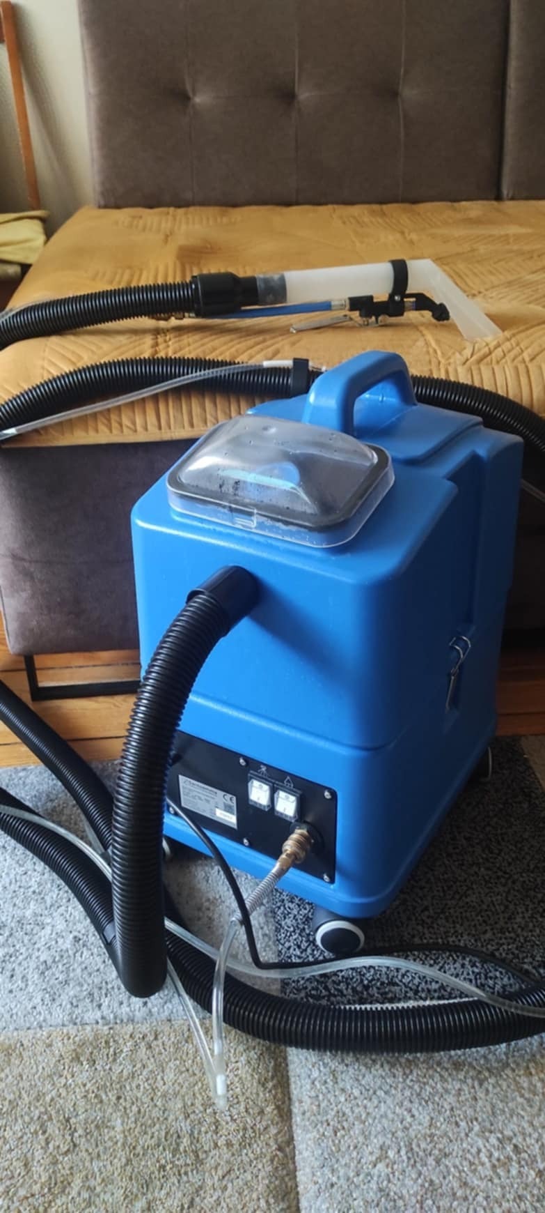 Niebieski odkurzacz piorący z przezroczystą pokrywą zbiornika, wąż i ssawka na tle brązowego zagłówka łóżka i beżowego dywanu.