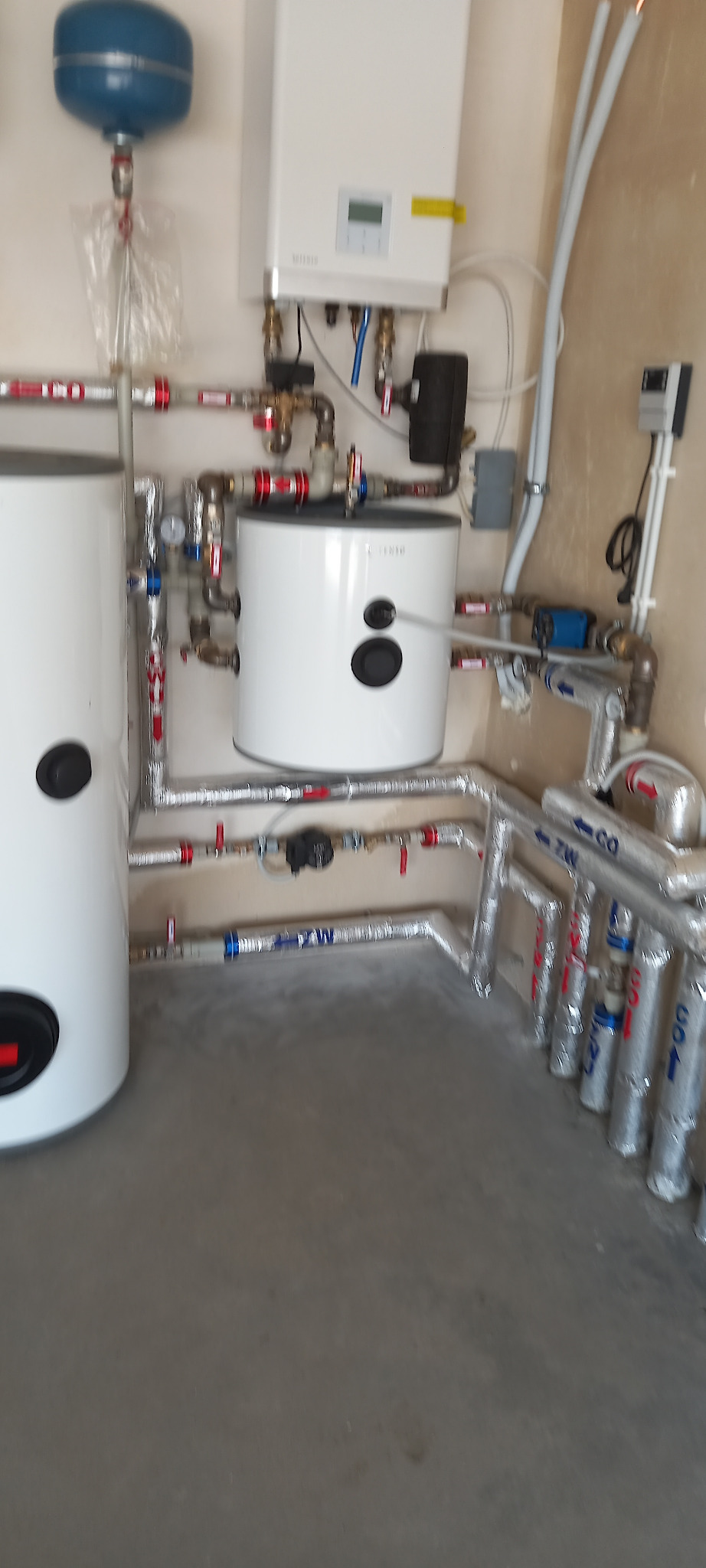 Biały piec gazowy z podłączonymi rurami w izolacji, zbiornik wyrównawczy i dodatkowe komponenty instalacji grzewczej na tle beżowej ściany.