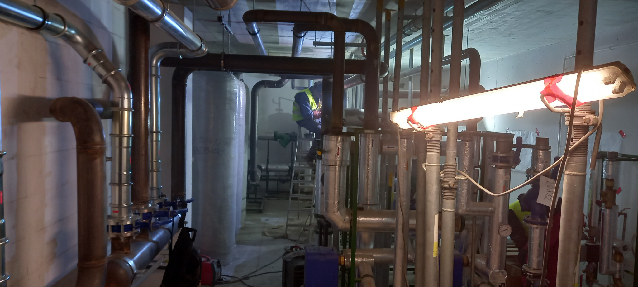 Instalacja grzewcza w kotłowni: widoczne liczne rury, zawory, zbiorniki, izolacja termiczna oraz pracownik w kamizelce odblaskowej na drabinie podczas montażu.