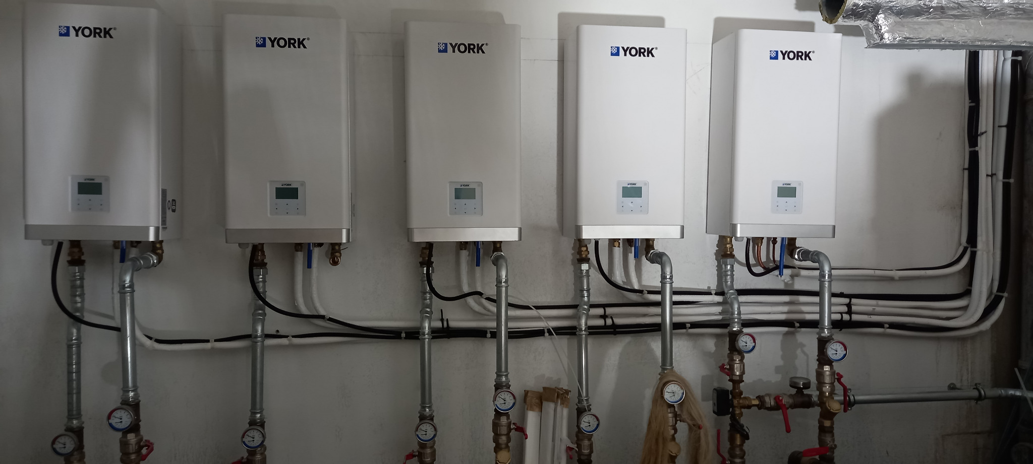 Rząd pięciu białych pieców gazowych marki York zamontowanych na ścianie, z widocznymi rurami i manometrami.