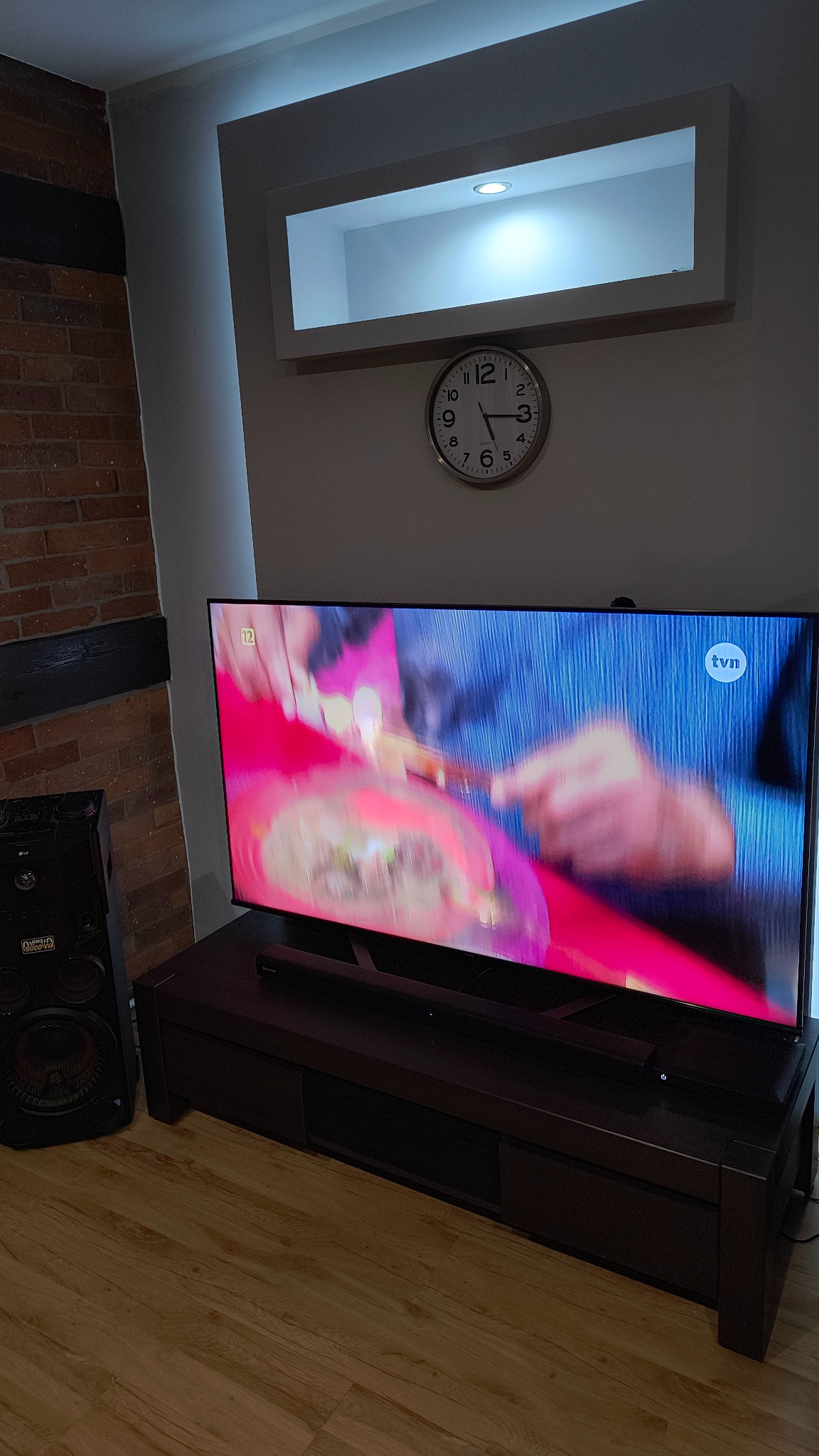 Telewizor na szafce RTV w salonie z ceglaną ścianą w tle, oświetlenie LED w suficie i zegar na ścianie.