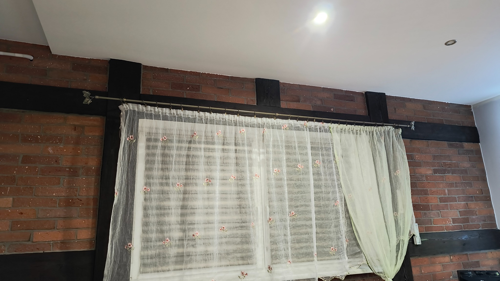 Fragment pokoju z odsłoniętym fragmentem ściany z czerwonej cegły, czarnymi belkami i oknem zasłoniętym białą, haftowaną firanką z motywem kwiatowym.