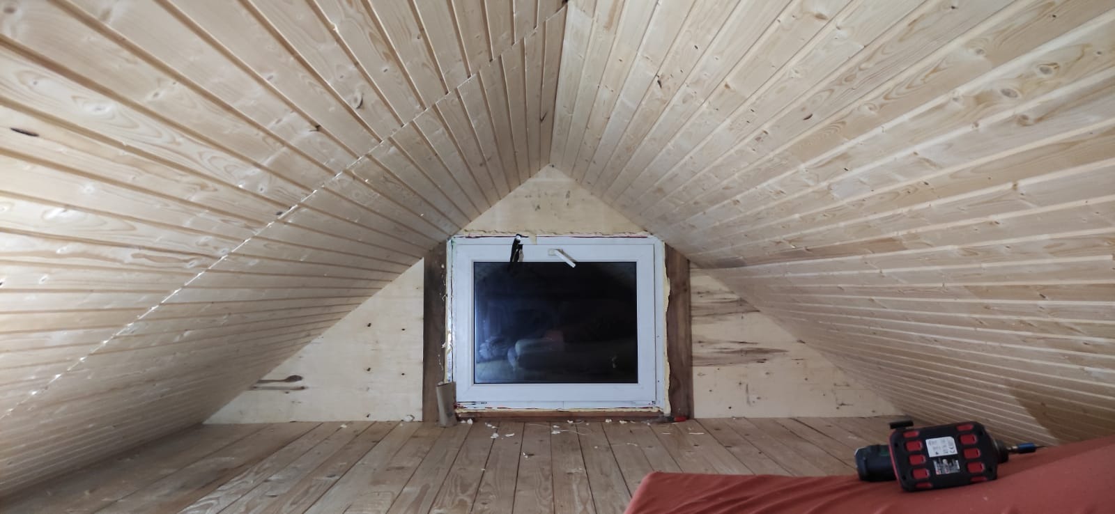 Poddasze z oknem w trakcie wykańczania drewnianą boazerią, widoczne narzędzie do montażu i fragment materaca.