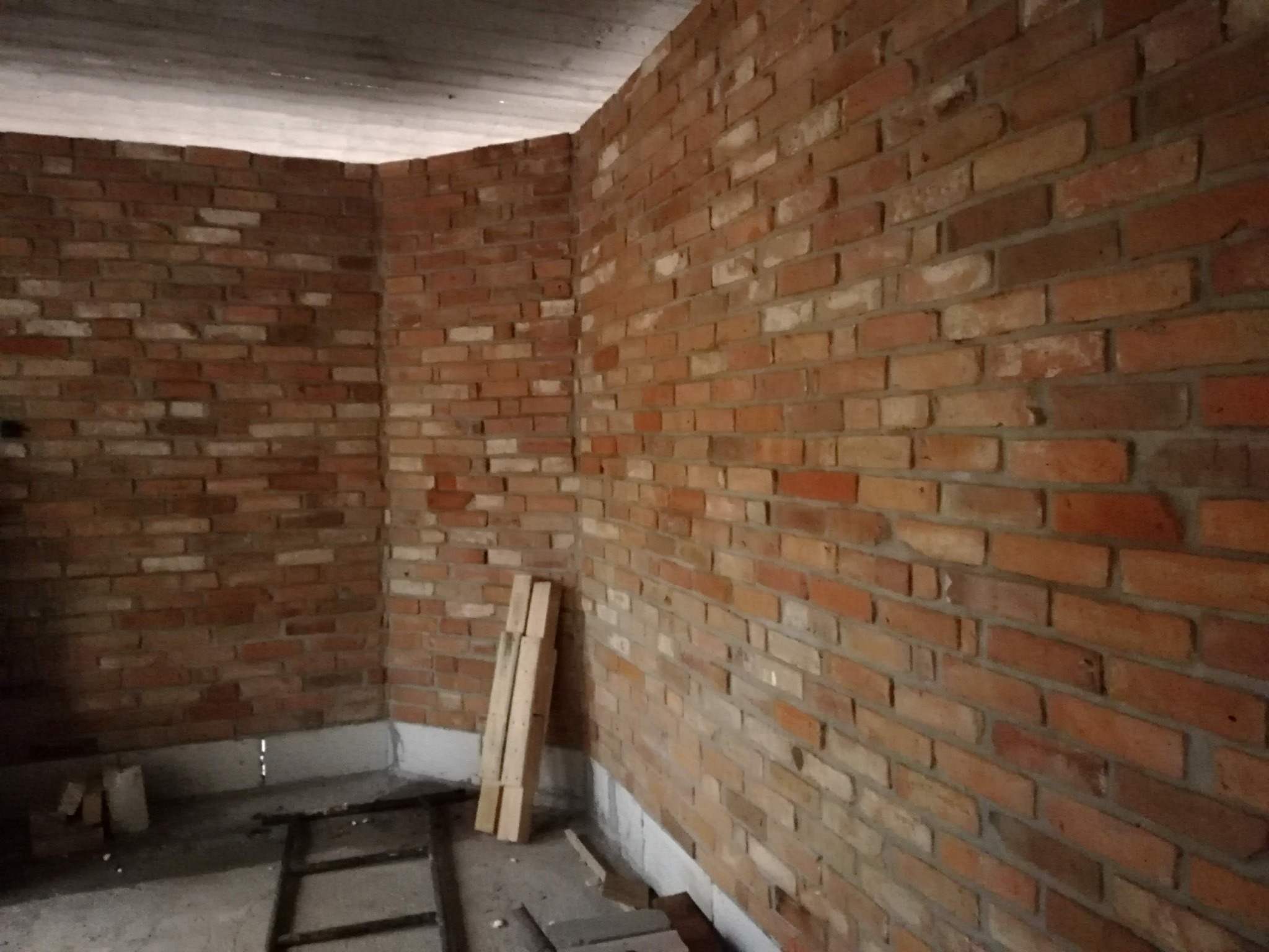 Surowa ściana z czerwonej cegły w nowo budowanym lub remontowanym pomieszczeniu, widoczne drewniane elementy na podłodze i surowy betonowy sufit.