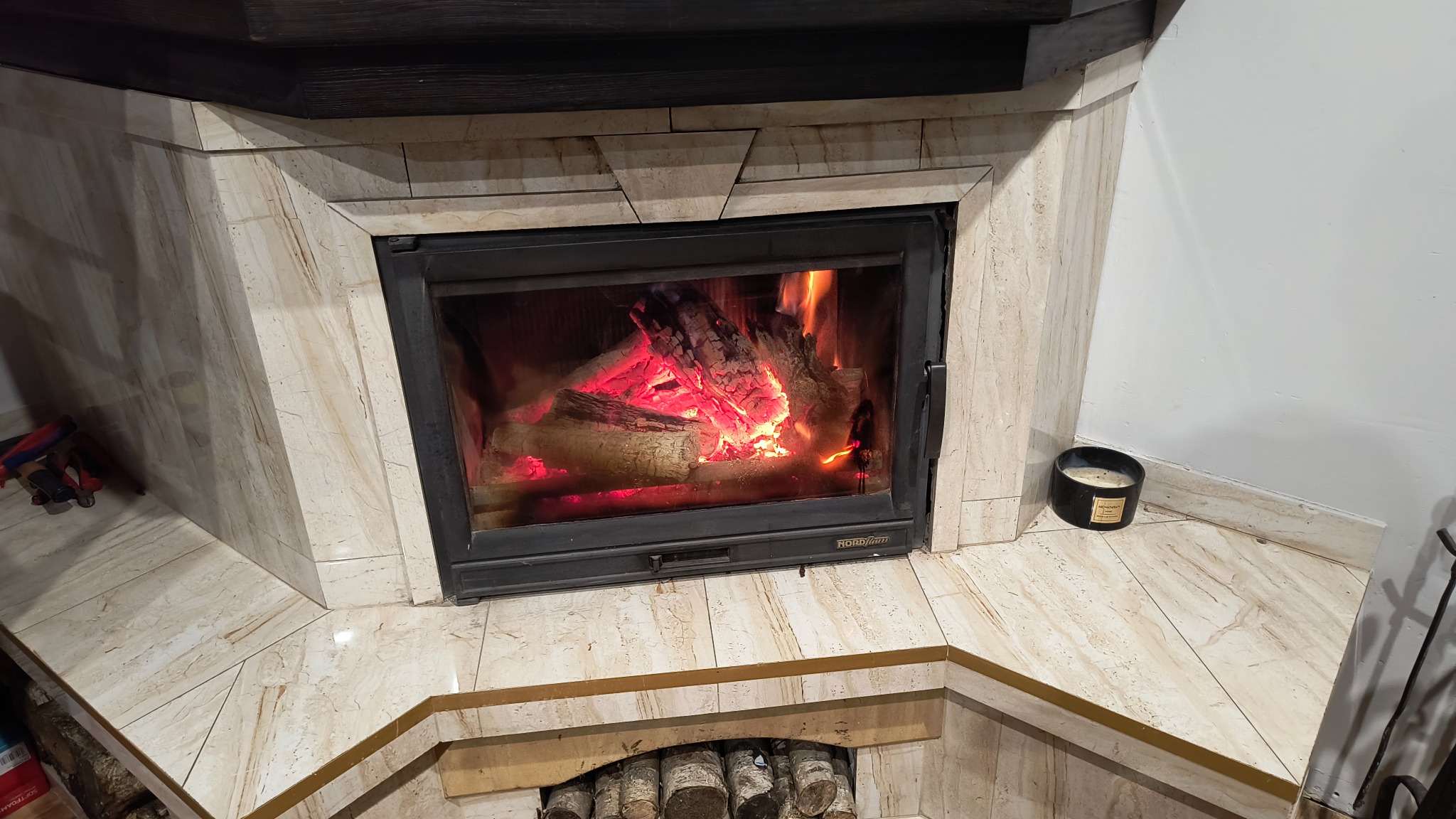 Obudowa kominka wykończona płytkami imitującymi marmur, z paleniskiem wypełnionym płonącymi polanami drewna i świecą zapachową obok.