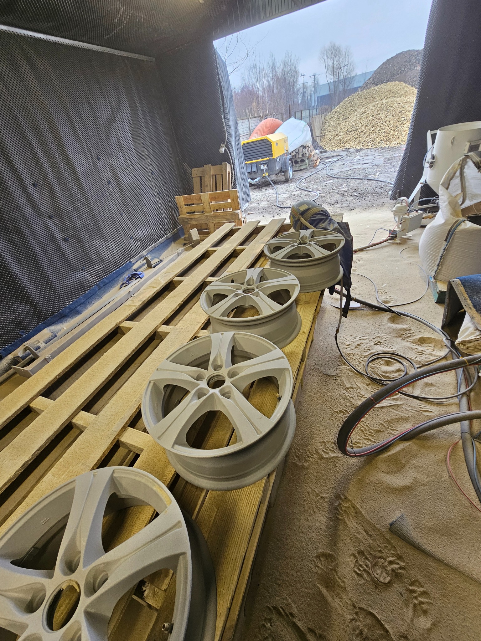 Szare felgi samochodowe ułożone na palecie w hali. W tle widoczny sprzęt i hałda piasku. Felgi po obróbce, przygotowane do dalszych prac.