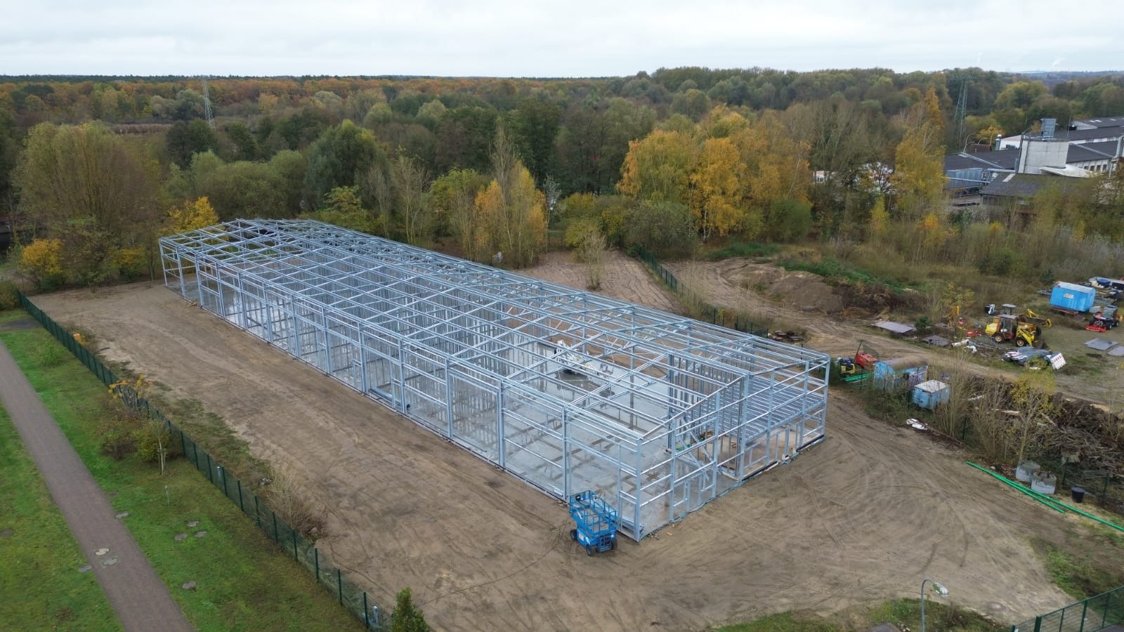 Szkielet hali stalowej w trakcie budowy, widok z góry, na ziemi widoczny podnośnik koszowy, w tle drzewa i budynki.