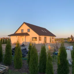 Dom jednorodzinny z ciemnym dachem w trakcie budowy, widoczne materiały budowlane, maszyna budowlana w tle i tuje na pierwszym planie w promieniach zachodzącego słońca.