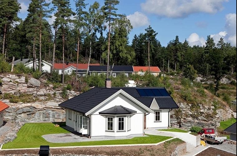 Nowoczesny, biały dom jednorodzinny z ciemnoszarą dachówką, panelami słonecznymi i charakterystycznym wykuszem okiennym, otoczony zielonym trawnikiem i naturalnym krajobrazem ze skałami i lasem w tle.