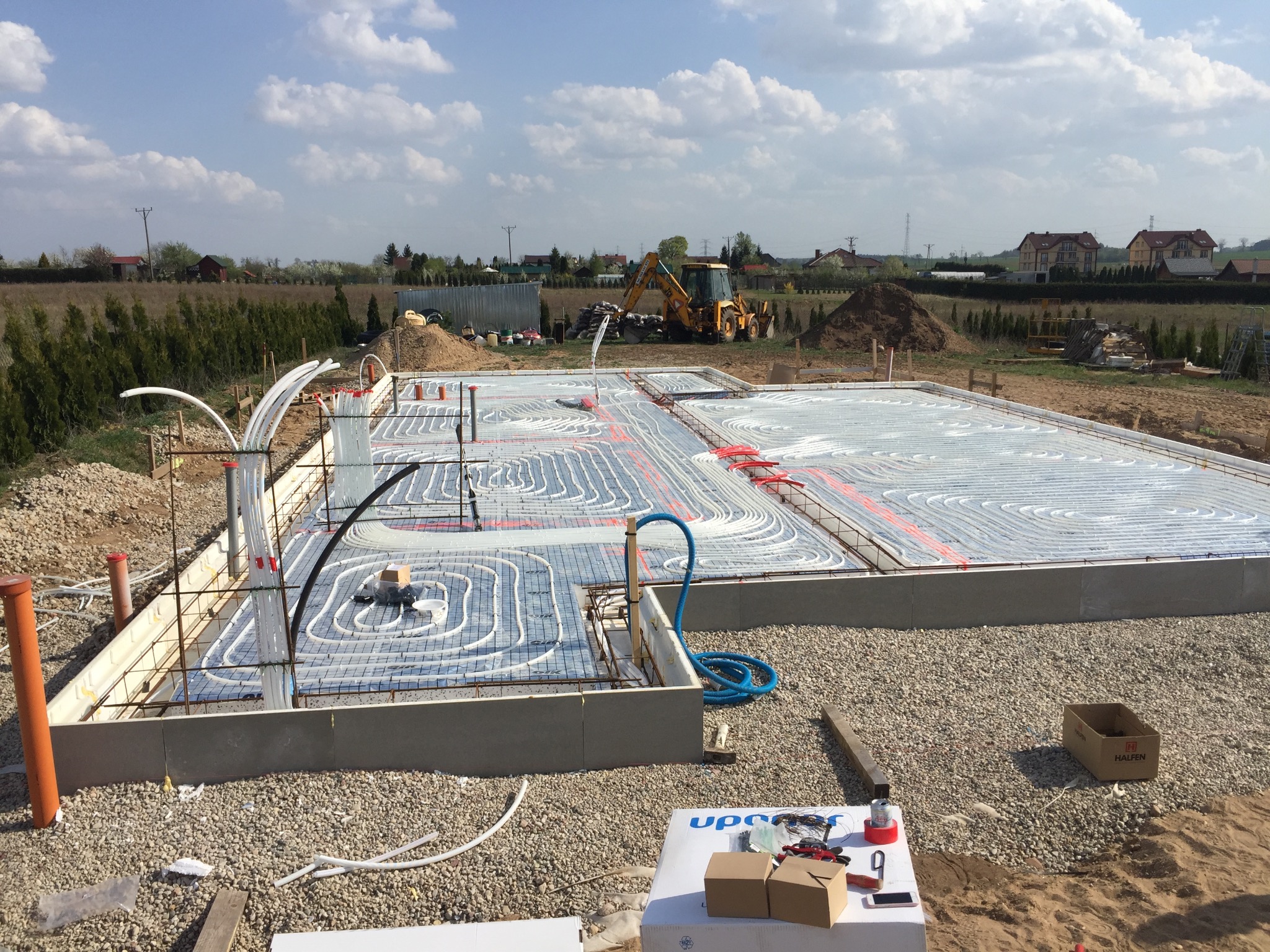 Fundament budynku z rozłożonym systemem ogrzewania podłogowego, widoczny także sprzęt budowlany i krajobraz wiejski w tle.