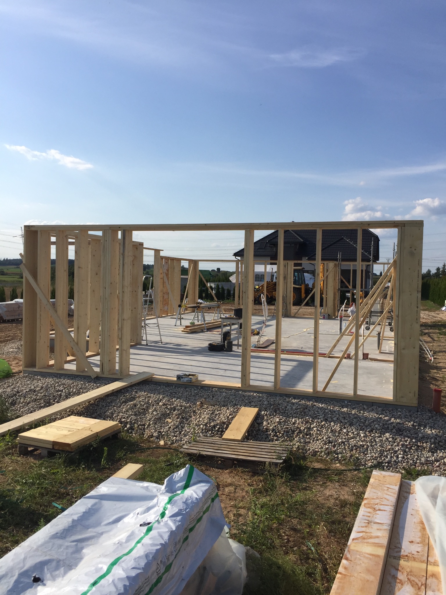 Wznoszenie szkieletu drewnianego budynku na betonowej płycie fundamentowej, widoczne z zewnątrz, w tle dom jednorodzinny i sprzęt budowlany.