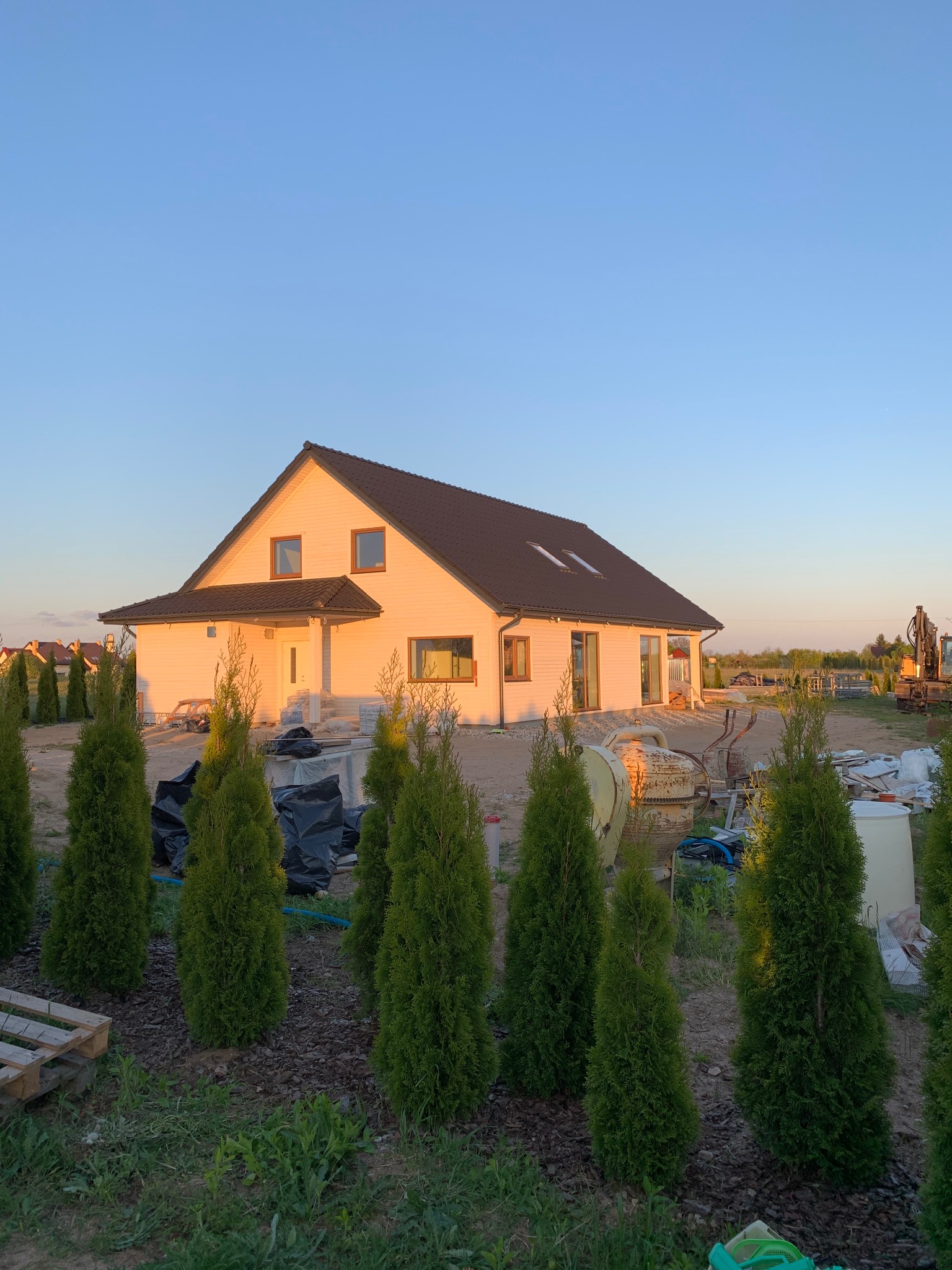 Dom jednorodzinny z ciemnym dachem w trakcie budowy, widoczne materiały budowlane, maszyna budowlana w tle i tuje na pierwszym planie w promieniach zachodzącego słońca.