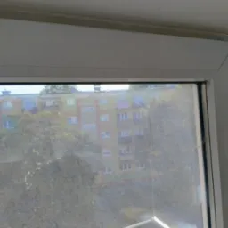 Brudna szyba okienna z widokiem na blok mieszkalny i drzewa, widoczne zacieki i osad na szkle.