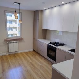 BOGDAN PYTEL Chem-Pol - Nowoczesna, jasna kuchnia w Radomiu po remoncie, z drewnianą podłogą, beżowymi szafkami i białymi górnymi szafkami, widok na okno z grzejnikiem.