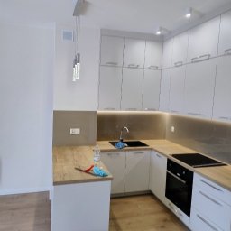 BOGDAN PYTEL Chem-Pol - Wnętrze nowoczesnej, jasnej kuchni z białymi szafkami i drewnianym blatem; na blacie leży ściereczka i butelka płynu, a na podłodze stoi czerwone wiadro.