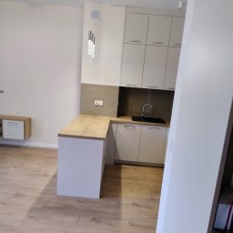 BOGDAN PYTEL Chem-Pol - Wnętrze jasnej, nowocześnie urządzonej kuchni z drewnianym blatem i białymi szafkami, widok na minimalistyczny zlew i baterię, a także wiszące lampy kryształowe.